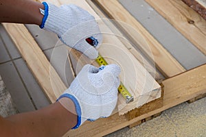 A man in white construction gloves measures a pine board with a tape measure