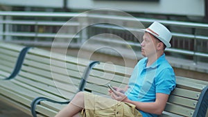 A man in a white hat is resting while sitting on a city bench, tracking a camera