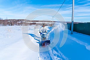 A man in a white jacket removes snow from a rural road with a blue snowblower in winter after a snowfall