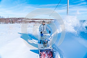 A man in a white jacket removes snow from a rural road with a blue snowblower in winter after a snowfall