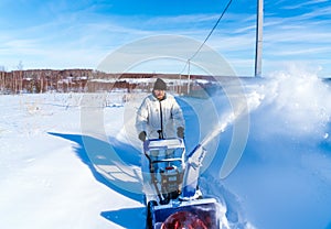 A man in a white jacket removes snow from a rural road with a blue snowblower in winter after a snowfall