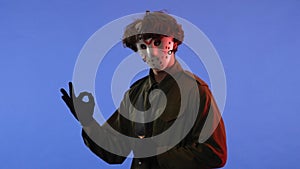 Man in white mask with sharp big knife in his hands showing OK gesture on blue background with red neon light. Image of