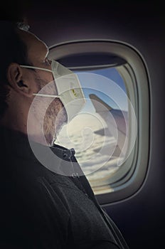 Man in a white medical mask looks thoughtfully at the clouds through the window of an aircraft