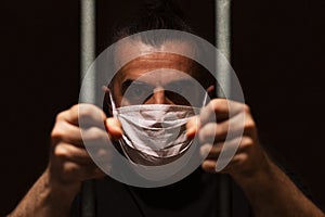 Man with white medical mask standing in the dark, holding bars with his bare hands, locked away in dark