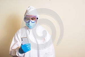 Man in a white protective suit, goggles and surgical gloves checks the cell phone during the coronavirus pandemic copy space