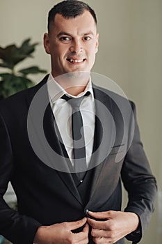 A man in a white shirt, black tie and black suit poses by the window and looks away. mends his jacket. Front view. A stylish watch