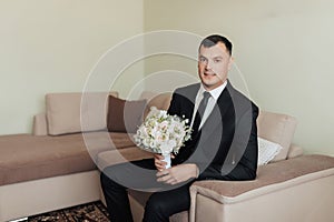 A man in a white shirt, black tie and black suit poses by the window and looks away. mends his jacket. Front view. A stylish watch