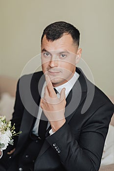 A man in a white shirt, black tie and black suit poses by the window and looks away. mends his jacket. Front view. A stylish watch