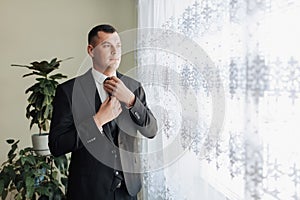 A man in a white shirt, black tie and black suit poses by the window and looks away. mends his jacket. Front view. A stylish watch