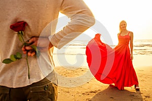 Man in a white shirt giving roses to surprised wonder woman in a long red dress on the ocean beach. romantic date