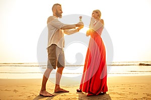 Man in a white shirt giving roses to surprised wonder woman in a long red dress on the ocean beach. romantic date