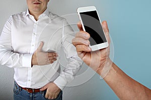 Man in a white shirt holds on to the chest in the region of the heart, next to his hand is holding a phone with black display, the