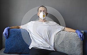 Man in white shirt, medical mask and rubber gloves is sitting on a sofa and feeling great staying home while pandemic