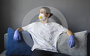 Man in white shirt, medical mask and rubber gloves is sitting on a sofa and feeling great staying home while pandemic