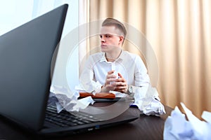 A man in a white shirt sits at a desk and crumples a paper in his hands. The concept of a financial crisis. The laptop