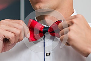 A man in a white shirt ties a red bow tie while preparing for a wedding ceremony