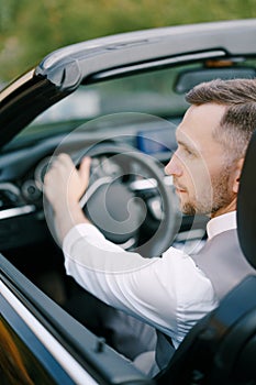 Man in a white shirt and vest sits at the wheel of a convertible. Back view. Close-up