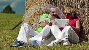 Man with white smartphone and woman with white tablet PC sits near a haystack