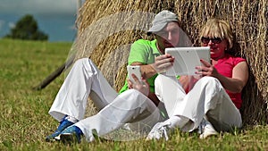 Man with white smartphone and woman with white Tablet-PC sits near haystack