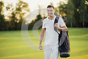 A man in a white suit walks around the golf course with a golf club bag and smiles