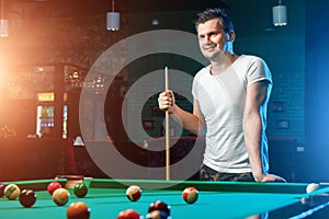 A man in a white T-shirt is playing billiards, a dark background. Pleasant pastime, entertainment, leisure, family holidays, games