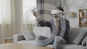 Man in white VR headset moves controllers and smiles playing online game and sitting on sofa against window slow motion