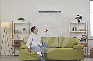 Man who's sitting on sofa at home turns on wall air conditioner using remote control