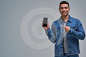 Man with a wide smile in a blue jacket holds a smartphone and points to it with the other hand - symbol from right to left.