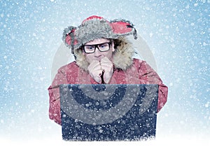 Man in winter clothes warming his hands while sitting in front of laptop. Cold, snow, blizzard