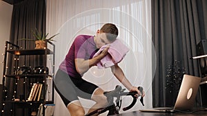 Man is wiping face with towel after hard cardio training on stationary bicycle. Male cyclist is cycling on indoor smart bike train