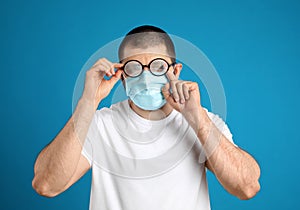 Man wiping foggy glasses caused by wearing disposable mask on blue background. Protective measure during coronavirus pandemic