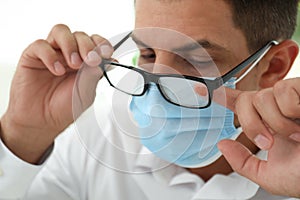 Man wiping foggy glasses caused by wearing medical mask indoors, closeup