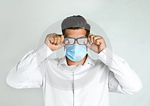 Man wiping foggy glasses caused by wearing medical mask on light background