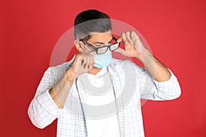 Man wiping foggy glasses caused by wearing medical mask on red background