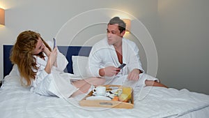 Man and woman in bathrobes making photos on smartphone eating in bed in hotel.