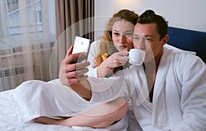 Man and woman in bathrobes making selfie photos on smartphone eating in hotel.