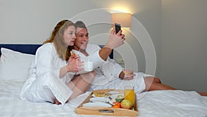 Man and woman in bathrobes making selfie photos on smartphone eating in hotel.