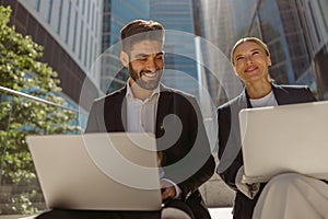 Man and woman in classic suit discussing business details and using laptop while sitting outdoors