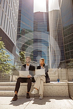 Man and woman in classic suit discussing business details and using laptop while sitting outdoors