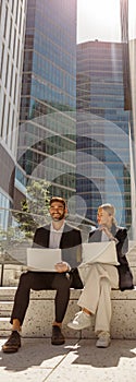 Man and woman in classic suit discussing business details and using laptop while sitting outdoors