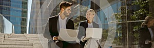 Man and woman in classic suit discussing business details and using laptop while sitting outdoors