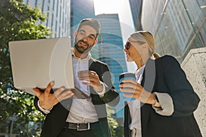 Man and woman in classic suit discussing business details and using laptop while standing outdoors