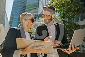 Man and woman in classic suit discussing business details and using laptop while standing outdoors