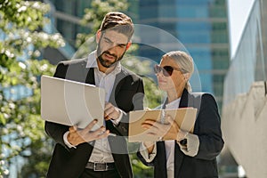 Man and woman in classic suit discussing business details and using laptop while standing outdoors