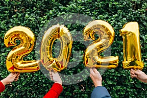 Man and woman couple hands holding gold foil balloons numeral 2021 in front of a wall of plants. New year celebration concept