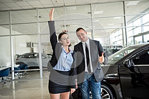 man and woman couple holding key to their car looking at camera and smiling in car showroom.