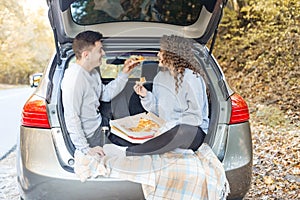A man and a woman are eating pizza near the car. Picnic in the fall