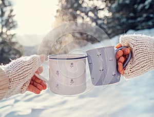 Man and woman hands in knitting mittens with cups of hot tea on winter forest glade