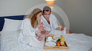 Man and woman having breakfast in bed in hotel and reading news in smartphone.