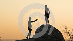 Man and woman hikers helping each other to climb from big stone at sunset in mountains. Couple climbing down high rock in evening